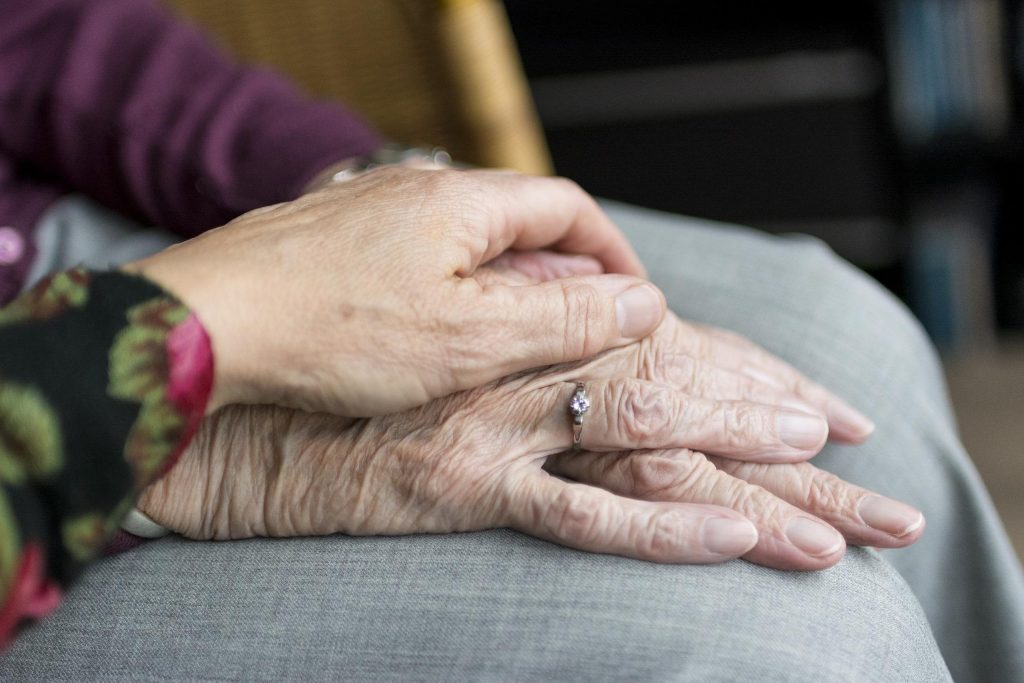 The hands of an elderly woman hold the hands of another woman, illustrating a tender moment of care and companionship.