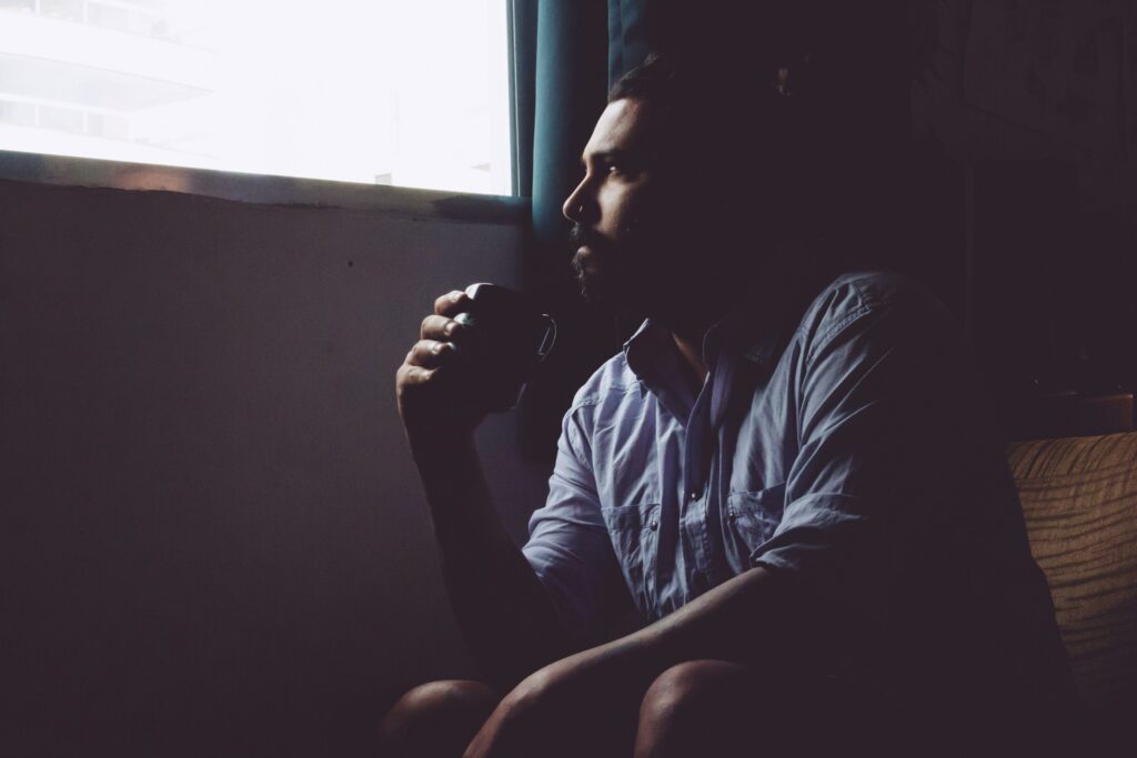 A man in a chair, holding a cup of coffee, looking out of the window in thought.