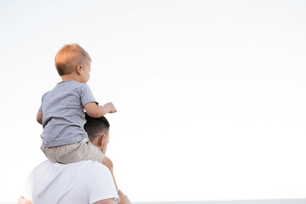 A father stands on the beach, carrying his young son on his shoulders.