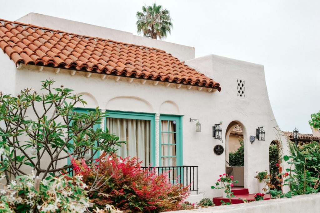 A white Spanish villa featuring a red tile roof and green trim, surrounded by lush greenery.