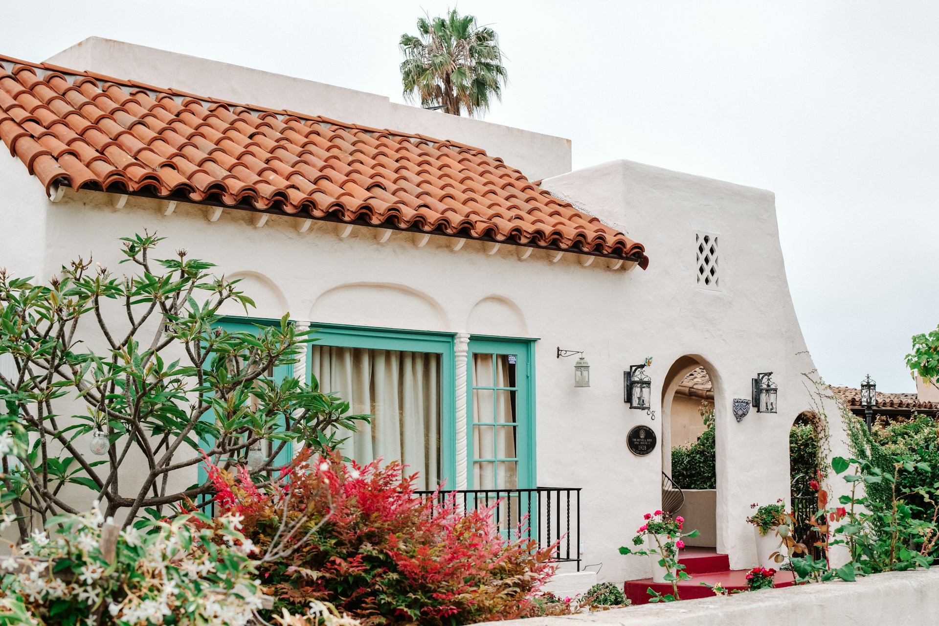 A white Spanish villa featuring a red tile roof and green trim, surrounded by lush greenery.